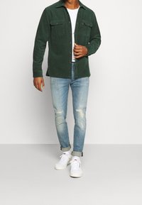Grön kordfljotskjorta med två bröstfickor, tillsammans med ljusblå slitna jeans och vita sneakers. Avslappnad passform och uppvikta ärmar.