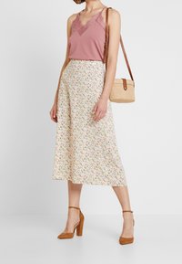 Ljusrosa linne med spetsdetaljer, tillsammans med en krämfärgad kjol med ett färgglatt blommönster. Modell som bär bruna klackskor med blockklack och en stråväska.