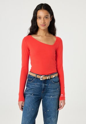 Vrouw met lang donker haar die een asymmetrische rode top met lange mouwen draagt, blauwe jeans met kleine hartpatronen en een beige riem.