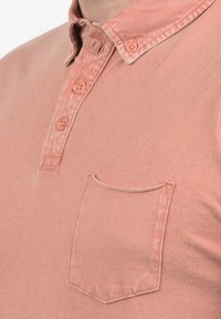 Svijetlo ružičasta polo majica od mekog materijala, s kopčanjem na gumbe, kontrastnim dugmadi i prednjim džepom zaobljenih rubova.