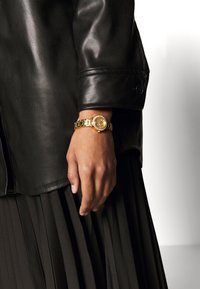 Reloj de oro con esfera redonda, pulsera de eslabones y acentos de cristal, llevado en una mano frente a una falda negra plisada y una chaqueta de cuero.