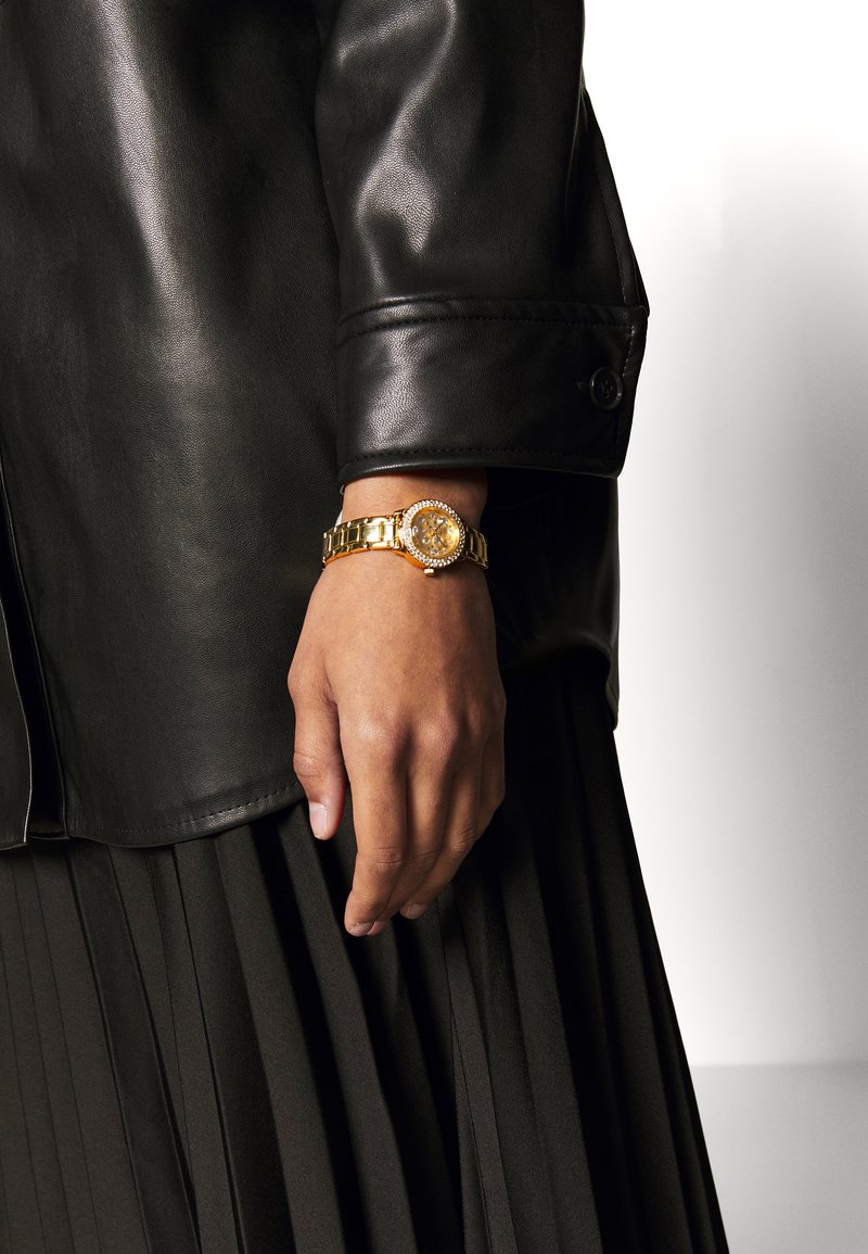 Gold watch with a round face, link bracelet, and crystal accents, worn on a hand in front of a pleated black skirt and leather jacket.