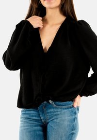 Blusa nera e tessurizzata con scollo a V profondo e maniche a palloncino, caratterizzata da una chiusura con bottoni davanti e un orlo annodato, abbinata a jeans blu chiaro.