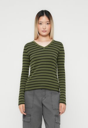 Woman wearing olive green and black striped long-sleeve V-neck top with gray cargo pants, standing against a plain white background.