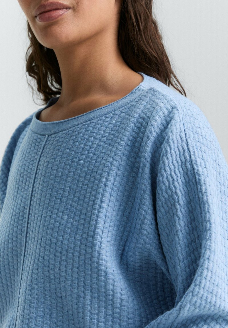 Jasnoniebieski sweter o fakturze z wzorem w gofry, szerokim dekolcie i opuszczonymi ramionami. Tkanina wydaje się miękka i przewiewna.