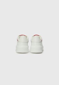 Weiße Sneakers mit einem glatten Lederoberteil, einer dicken Sohle und roten Akzenten an der Ferse, mit rotem Branding an den Seiten.