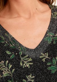 Top nero a scollo a V con una texture scintillante caratterizzata da motivi di foglie verdi e accenti floreali bianchi. Dettaglio ravvicinato del collo e delle trame del tessuto.