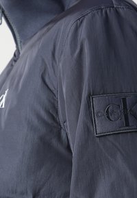 Kurtka Calvin Klein z ciemnoszarym nylonowym zewnętrzem, wysokim kołnierzem oraz czarną tkaninową naszywką z logo na lewym rękawie.