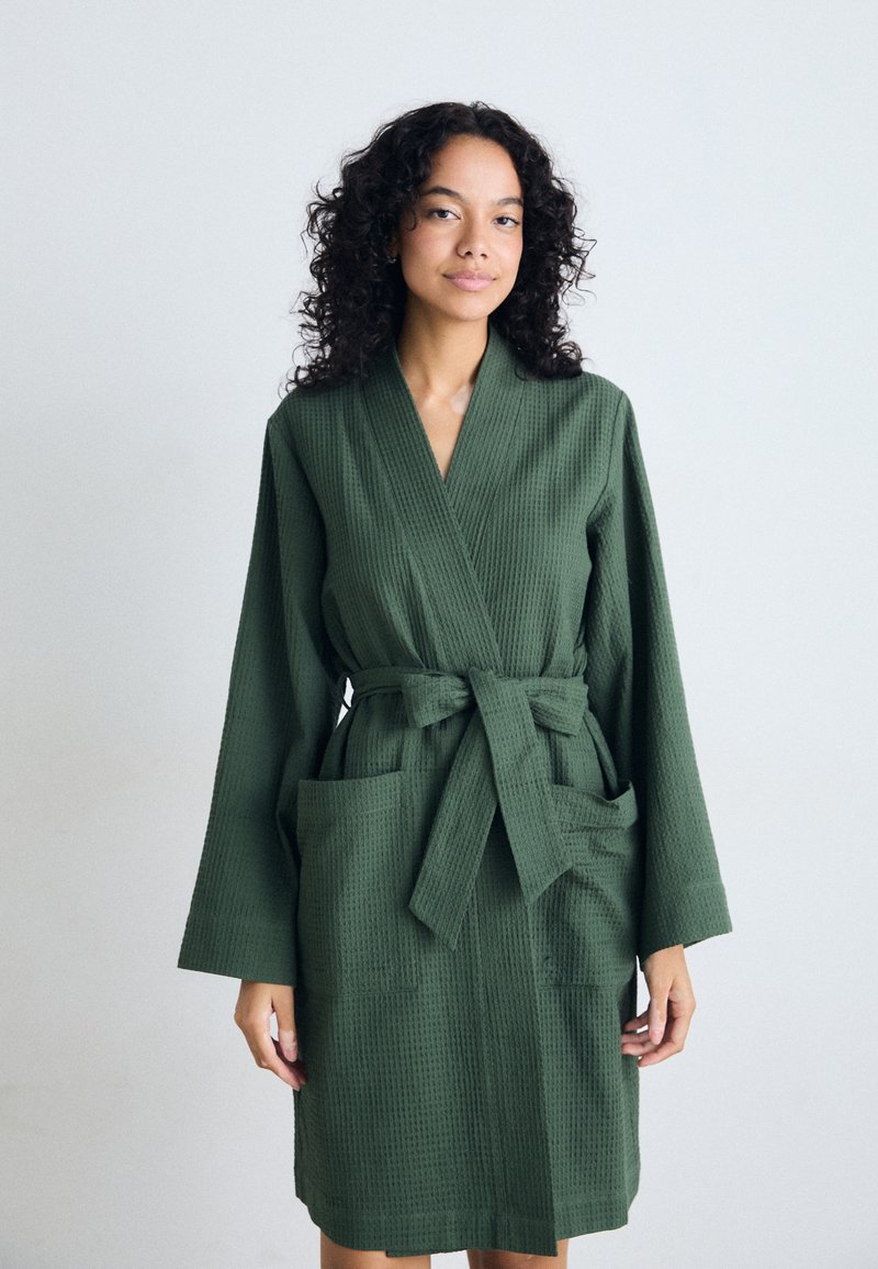 Robe vert à texture gaufrée avec des manches longues, une ceinture à nouer et deux poches avant. Le tissu semble doux et léger.