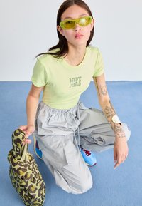 Tricou Nike verde deschis, pantaloni gri cu șnur și adidași albastre. Accesoriile includ ochelari de soare galbeni și o geantă verde cu model.