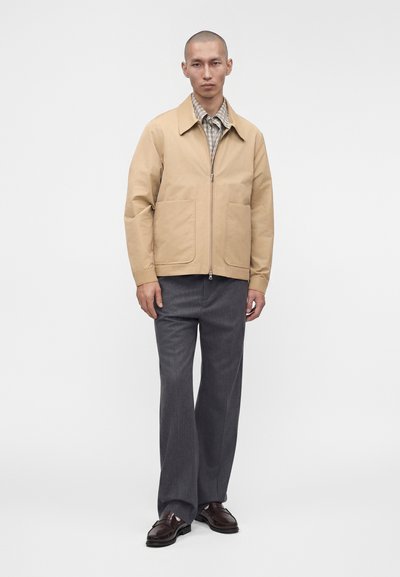 Homme portant une veste beige zippée, une chemise à carreaux, un pantalon gris et des mocassins marron foncé, debout devant un fond blanc uni.