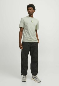 Camiseta verde claro con un logo cuadrado en el pecho, combinada con pantalones negros holgados y zapatillas beige con acentos grises.