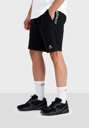 Shorts sportivi neri con tasche laterali, caratterizzati dal testo bianco "LE COQ SPORTIF". Abbinati a calze bianche a crew e sneakers nere.