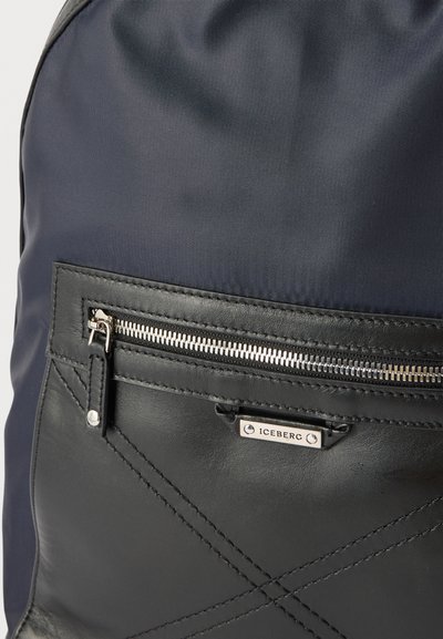 Μπροστινή τσέπη μαύρου και ναυτικού μπλε σακιδίου με ασημένιο φερμουάρ και μικρή μεταλλική ετικέτα με την επιγραφή "ICEBERG" κεντραρισμένη κάτω από το φερμουάρ.