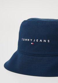 Námornícky modrý vedierkový klobúk z bavlny s vyšívaným logom "TOMMYJEANS" a malým farebným emblémom. Má široký, smerom nadol sklonený okraj.