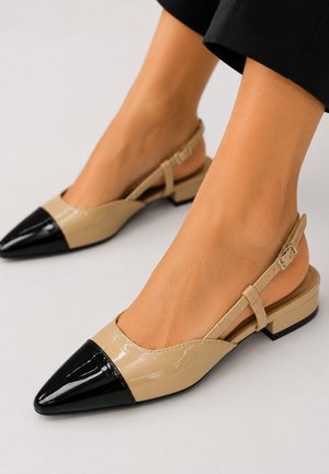 Vrouw die beige en zwarte lakleren slingback platte schoenen draagt met lage blokhakken en puntige neuzen, afgebeeld op een lichte achtergrond.