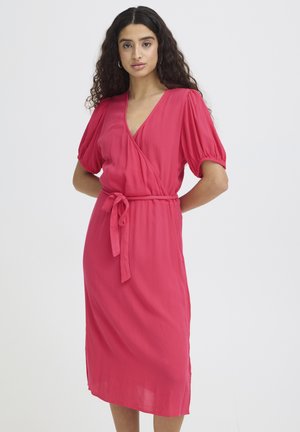 Femme aux longs cheveux bouclés portant une robe portefeuille rose mi-longue avec des manches bouffantes et une ceinture nouée à la taille, debout devant un fond blanc.