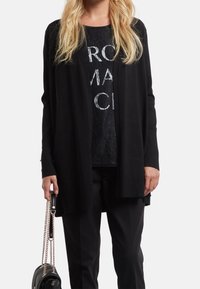 Cardigan lungo nero realizzato in morbido tessuto, con apertura frontale, abbinato a un top in pizzo nero con audace lettering bianco e pantaloni neri. Accessori: borsa nera piccola.