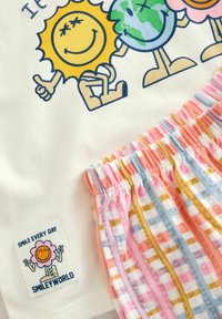 Personaggi animati di sole e terra che si tengono per mano su tessuto bianco, con una fascia elastica a righe colorate sotto e una toppa con la scritta "Sorridi ogni giorno".