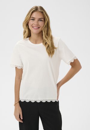 Mladá žena s dlhými blond vlasmi má na sebe biele tričko s krátkym rukávom a čipkovaným lemom a čierne nohavice, usmieva sa na jednoduchom pozadí.