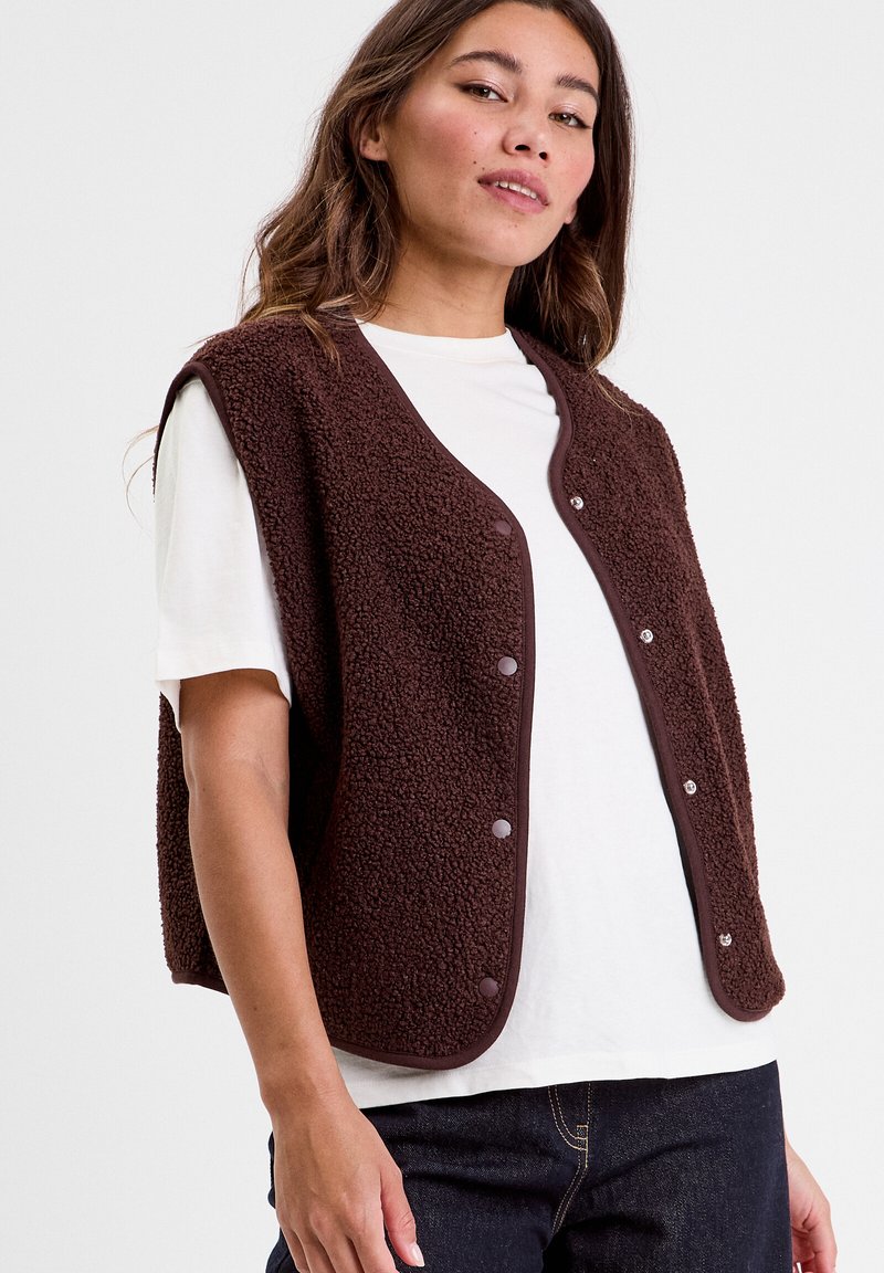 Brown, textured vest with snap buttons, round neckline, and sleeveless design, worn over a white shirt and dark jeans.