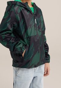 Černá a zelená větrovka s kapucí, zapínáním na zip a bočními kapsami. Má lehkou texturovanou tkaninu.