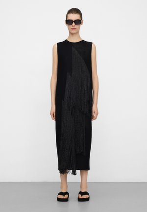 Schwarzes ärmelloses Kleid mit strukturierter Fransenverzierung auf einer Seite, knielang, kombiniert mit Plateausandalen. Glatter Stoff, minimalistisches Design.