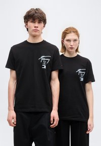 Νέος άνδρας και γυναίκα στέκονται δίπλα δίπλα φορώντας μαύρα T-shirts με σχέδια, με τα ονόματα πόλεων τυπωμένα στο στήθος, σε απλό λευκό φόντο.