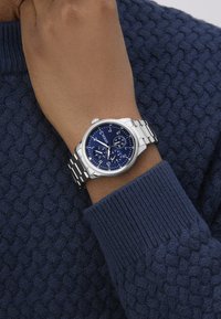 Ezüst fém szíjjal rendelkező karóra, kék számlappal és több alszámlappal, egy texturált sötétkék pulóver ujján viselve.