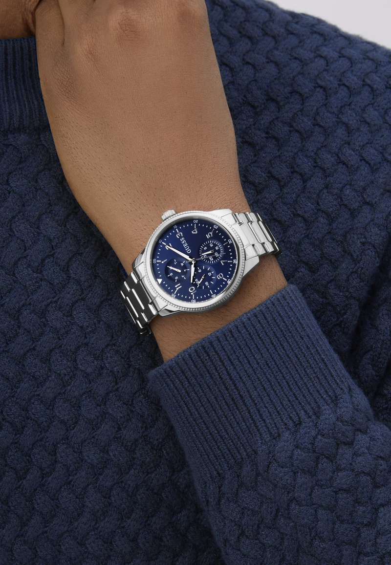 Ezüst fém szíjjal rendelkező karóra, kék számlappal és több alszámlappal, egy texturált sötétkék pulóver ujján viselve.