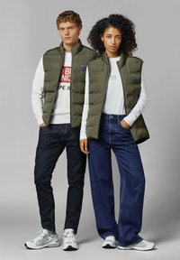 Olijfgroene puffer vesten met ritssluitingen, gedragen over witte tops, gecombineerd met donkere jeans en sneakers. Gladde textuur en geborduurd ontwerp.