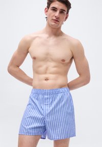 Jeune homme aux cheveux courts et foncés, portant un boxer à rayures bleues et blanches, debout avec les mains croisées dans le dos sur un fond blanc uni.