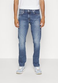 Jeans de denim azul claro con corte recto, detalles desgastados, cinco bolsillos y botón de metal. Combinados con zapatillas grises y azules.