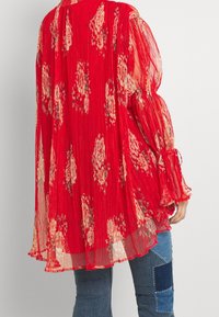 Punainen laskostettu pusero, jossa on kukkakuosi, ilmava muotoilu, pitkät hihat, joissa on nauhakoristeet, yhdistetty sinisiin kirjailtuihin farkkuihin.