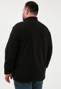 Chaqueta de forro polar negra con un cuello alto, mangas largas y una textura suave. La vista trasera muestra un corte relajado y sin cierres visibles.