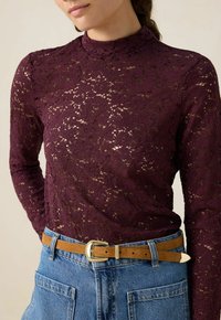 Burgunderfarbenes Spitzen-Oberteil mit langen Ärmeln und floralem Muster, hohem Kragen, kombiniert mit blauen Denim-Shorts und einem braunen Gürtel mit goldener Schnalle.