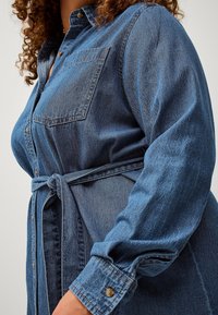 Abito camicia in denim blu scuro con chiusura frontale a bottoni, tasca sul petto, maniche lunghe e cintura annodata in vita per una silhouette aderente.