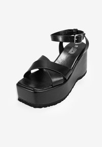 Sandalia de plataforma negra con una amplia tira frontal, tiras cruzadas en el tobillo y un broche. Material sintético suave, tacón grueso y suela texturizada.