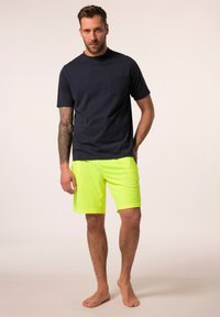 Námorně modré tričko s krátkým rukávem a kapsou na prsou, spárované s neonově žlutými šortkami. Hladká textura, volný střih. Model stojí bos.