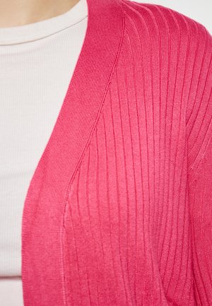 Pletený, ribovaný cardigan v jasně růžové barvě, s výstřihem do V a jemnou texturou, kombinovaný s bílým přiléhavým topem pod ním.