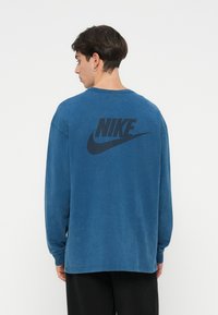 Plava pamučna majica dugih rukava s velikim crnim Nike logotipom na leđima, s opuštenim krojem i rebrastim manžetama.