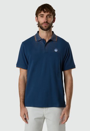 Uomo con barba che indossa una polo blu navy con rifiniture arancioni e pantaloni bianchi, in piedi davanti a uno sfondo chiaro e uniforme.