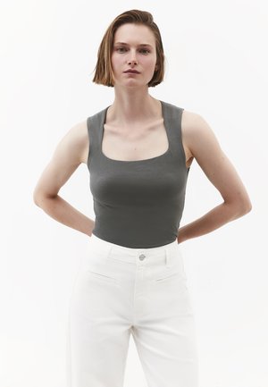 Mujer con cabello corto y castaño que lleva una camiseta sin mangas gris oscuro y pantalones blancos de talle alto, de pie con las manos detrás de la espalda.