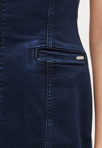 Džínové šaty tmavomodrej farby s priliehavým strihom. Majú bočné vrecko s kovovou značkou a viditeľné detaily prešívania. Mäkká textúra.