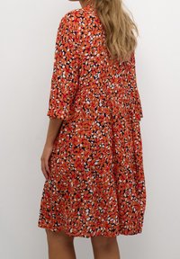 Červené květinové šaty s tříčtvrtečními rukávy, které mají vrstvený design. Látka má jemnou texturu s živým multicolor vzorem.