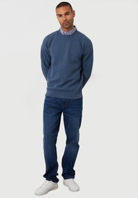 Blauwe gestructureerde trui met ronde hals, gecombineerd met donkerblauwe jeans en witte sneakers. De outfit heeft een laag onder een lichtblauw shirt.