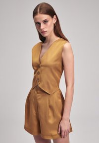Conjunto de dos piezas en mostaza con un top corto sin mangas con botones y pantalones cortos de tiro alto y plisados. Textura suave, corte ajustado.