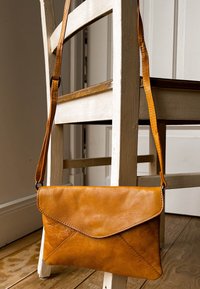 Tan leren crossbody tas met een envelop ontwerp, voorzien van een gestructureerd oppervlak, een verstelbare schouderband en zichtbare stikdetails.