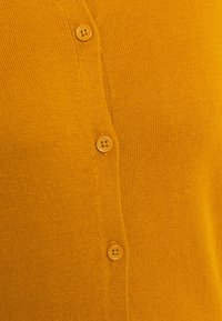 Gros plan sur un cardigan en maille jaune moutarde avec quatre boutons assortis fixés au centre.