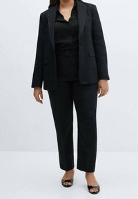 Blazer nero su misura e pantaloni coordinati, realizzati in tessuto leggero. Include una camicia aderente e sandali neri con punta aperta.